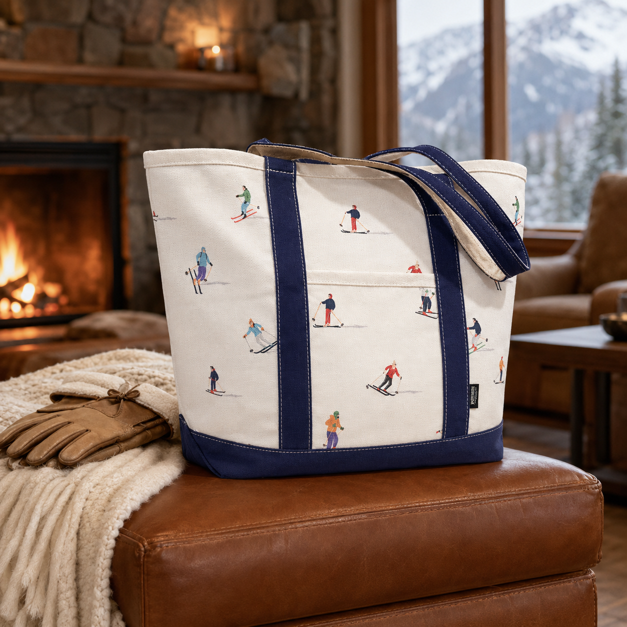 A canvas tote bag with a navy blue trim and handles, featuring a repeating pattern of skiers on an off-white background, sitting on a brown leather ottoman in a cozy living room setting with a fireplace and mountain view.