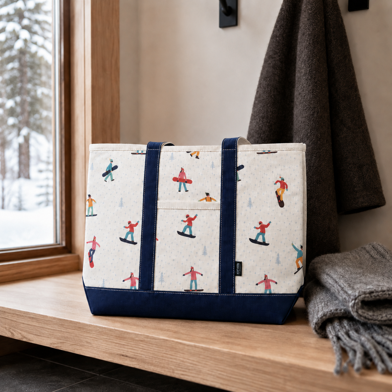 A canvas tote bag with a colorful skier pattern, featuring navy blue straps and bottom trim, resting on a wooden windowsill next to a gray blanket.