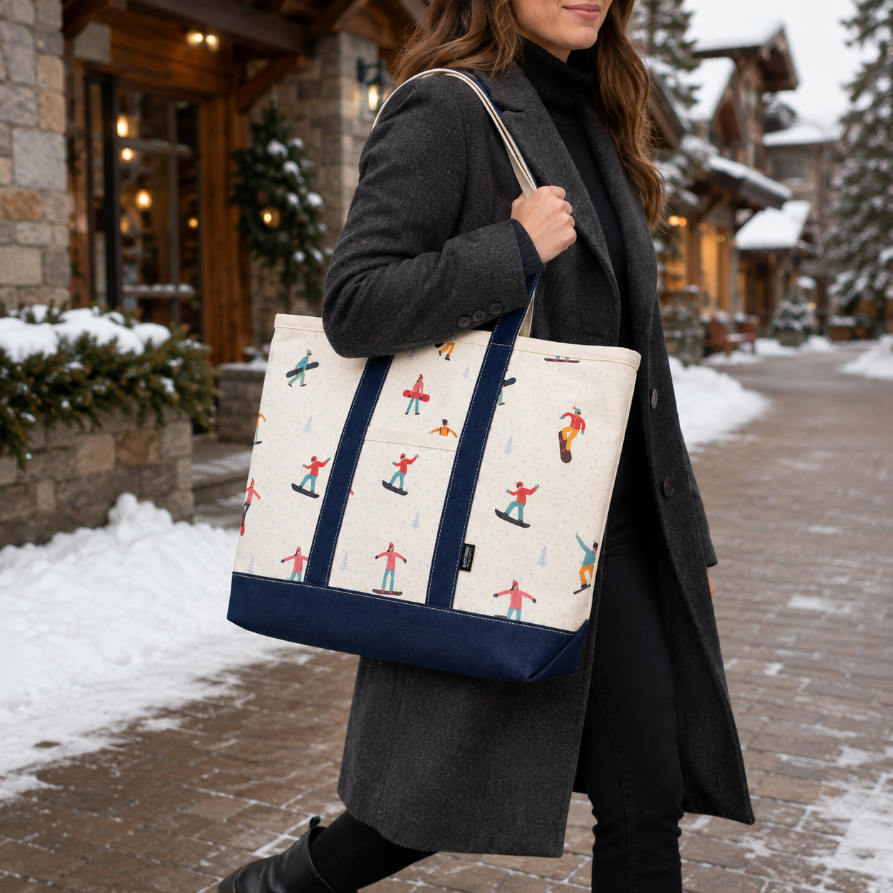 A woman in a dark gray coat carries a large canvas tote bag with a cream background and a colorful print of skiers and snowboarders, featuring navy blue straps and bottom panel, walking along a snowy outdoor path.