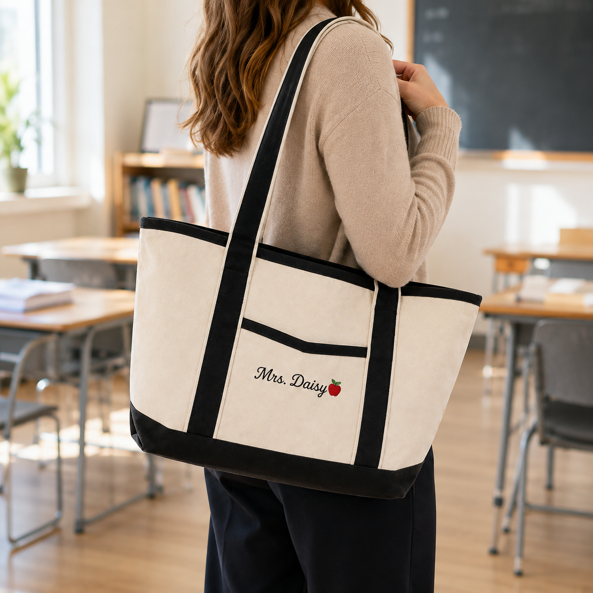 A woman in a beige sweater carries a large cream canvas tote bag with black trim and handles in a bright classroom. The bag features a front pocket with the embroidered text 'Mrs.