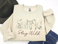 Stone colored sweatshirt with wildflowers embroidered onto the front of the sweatshirt with the words stay wild delicately embroidered underneath the wild flowers.