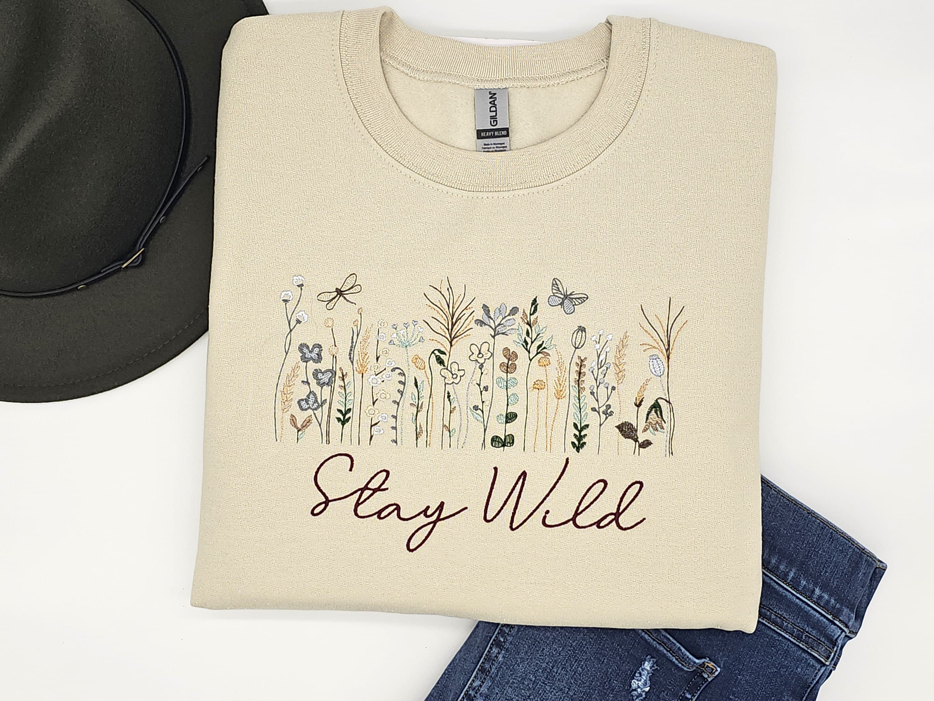 Stone colored sweatshirt with wildflowers embroidered onto the front of the sweatshirt with the words stay wild delicately embroidered underneath the wild flowers.