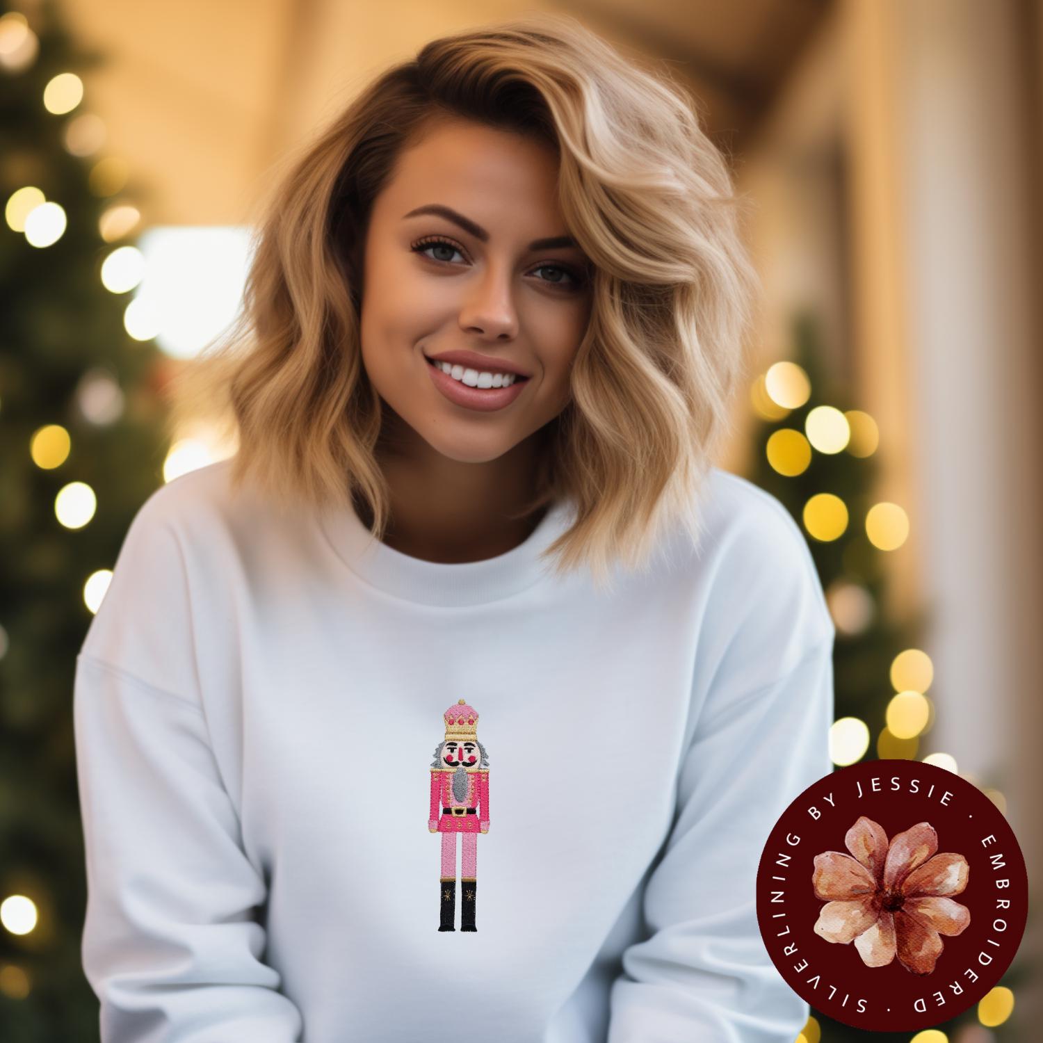 White crewneck fleece lined sweatshirt with a pink nutcracker embroidered onto the front.